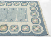 A refined mid-century Swedish flat weave by celebrated designer Ingegerd Silow, signed with her "IS" monogram in the lower right corner. Woven in soft glacier blues, cream, rose, and sage, this piece embodies the quiet elegance and warmth that define Scandinavian Modern textile design.

Silow’s signature geometric floral medallions create a rhythmic border around a subtly striated blue field, giving the rug both structure and softness. Handwoven in wool on a linen foundation, the texture is supple yet sturd