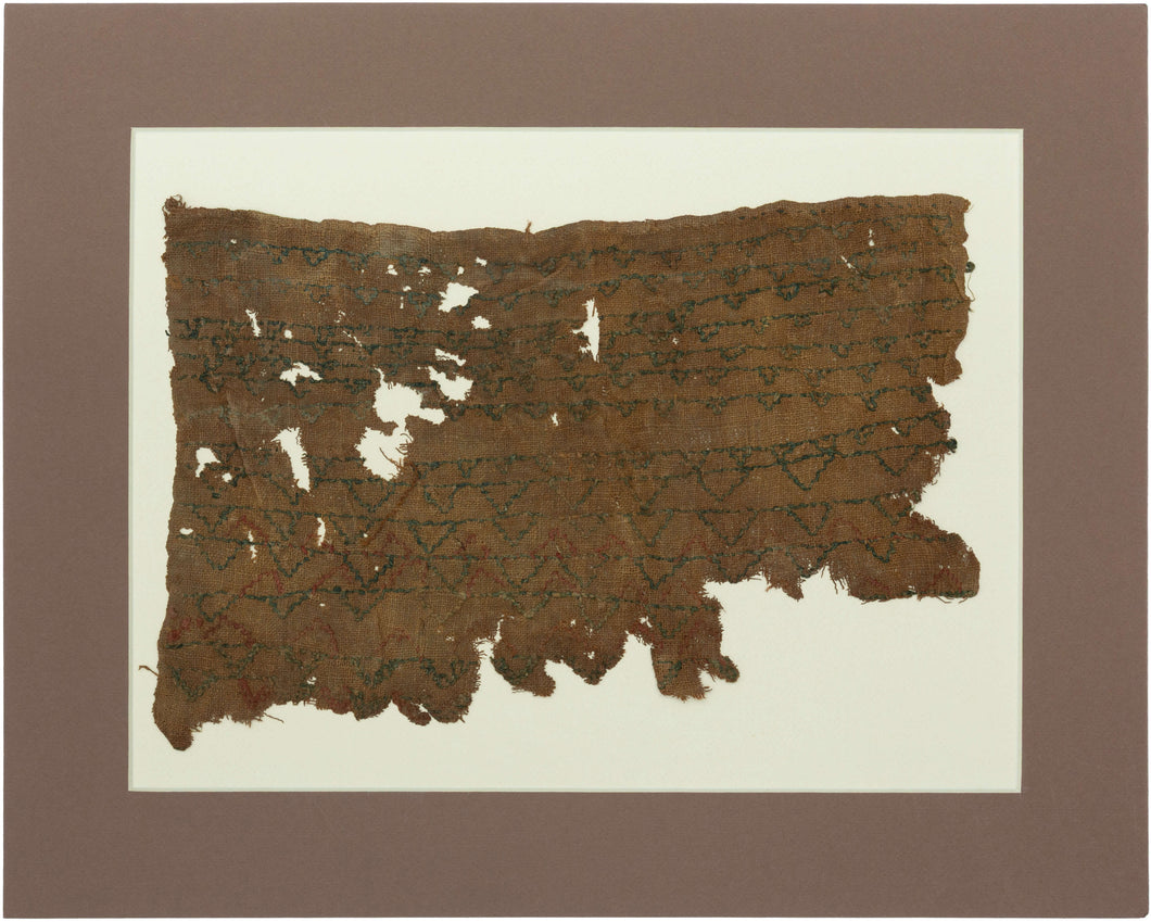 A remarkably understated Coptic textile fragment, woven on a warm brown linen ground and patterned with repeated horizontal bands of small linked motifs in deep blue-green, with traces of red.

Rather than the bold tapestry panels more often associated with Coptic textiles, this piece has an appealingly spare, almost minimalist quality. The repeating forms create a quiet rhythm across the surface, their irregularities revealing the hand of the weaver.