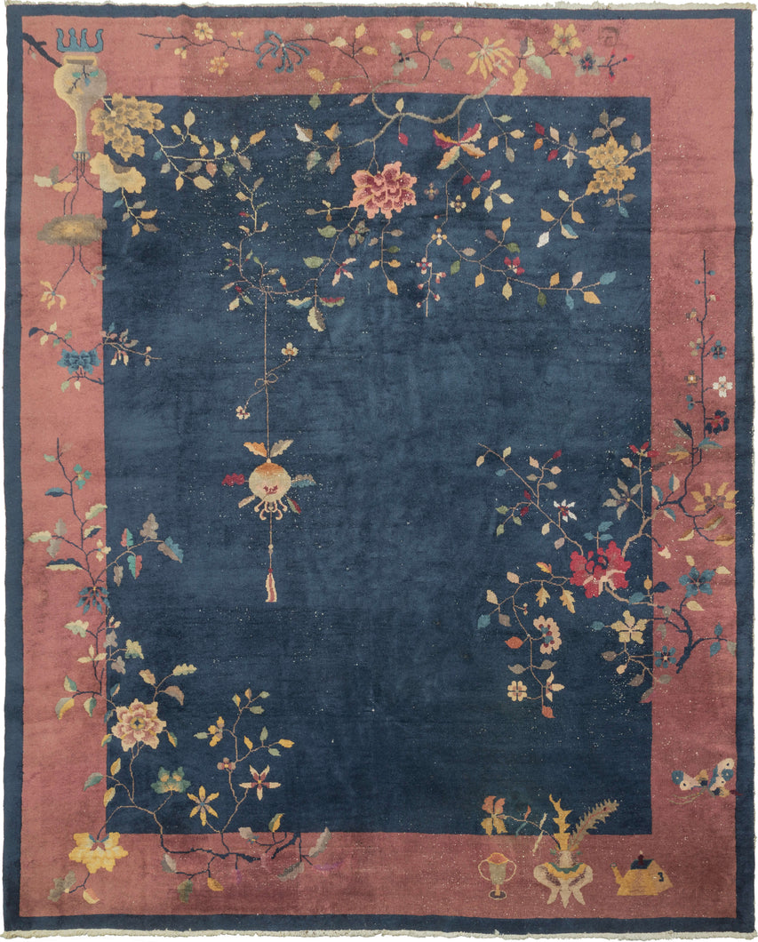 This Deco rug was handwoven in Tietsin, China during the second quarter of the 20th century.   This stately deco features a navy ground with a calm mauve border. The design of blossoming branches and potted plants is whimsical and not restrained by the border.  A single lantern hangs near the center of the rendering.