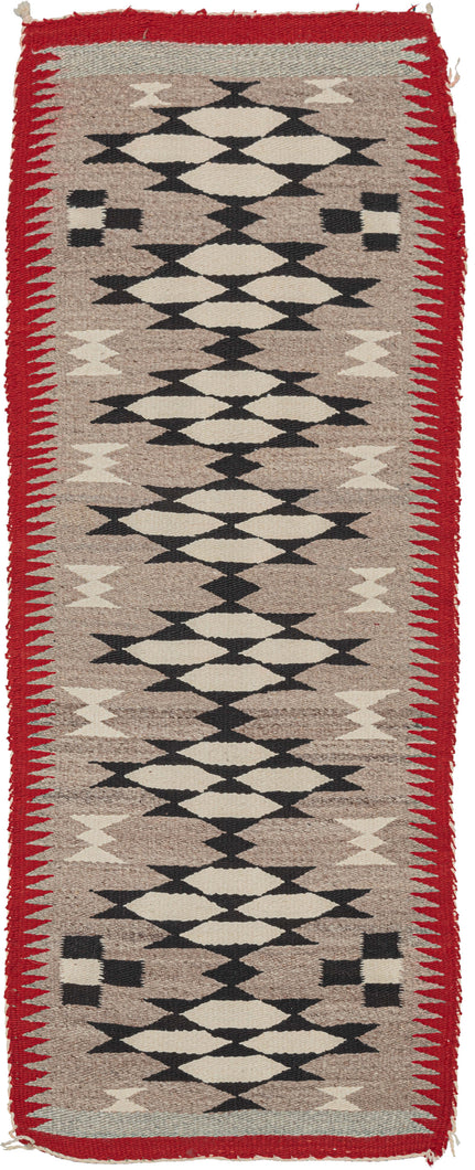 This Navajo rug was handwoven in the Southwest US during the second quarter of the 20th century.  It features interconnected rosette-like diamonds in black and white on a vast gray field. The gray color is created by mixing different strands of undyed wool. The whole is framed by a thin red border that sawtooths into the gray field.