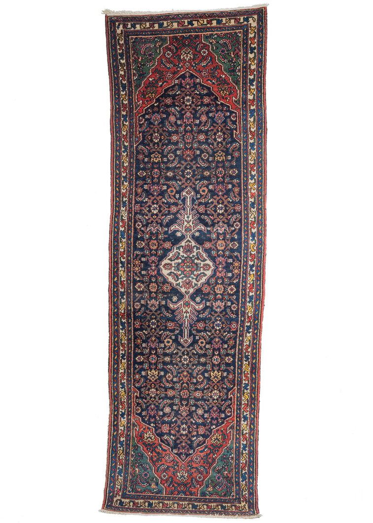 Bibikabad runner featuring a small but prominent ivory medallion with a single Herati motif atop an allover Herati field in deep indigo. The field cuts in at both ends giving way to first red then green backdrops. The border is composed of scrolling vines with pops of yellow, green red, pink and blue.