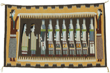A finely woven Yei Bi Chei pictorial by celebrated Diné weaver Della Woody Begay, depicting a ceremonial procession of masked figures associated with the Navajo Nightway tradition. The dancers are arranged in a rhythmic line between two larger attendant figures, set against a deep brown ground and framed by Woody’s particularly distinctive serrated, cloud-like border.