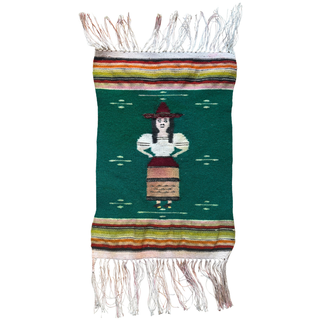 A charming mid–20th century Zapotec weaving from Oaxaca, Mexico, depicting a standing female figure in traditional dress. She wears a wide-brimmed red hat, a white blouse with puffed sleeves, and a striped skirt rendered in earthy tones. Her bold black hair and expressive features are woven with striking clarity, giving the figure a distinctive presence. Metal thread has been utilized in accents of the hat.

