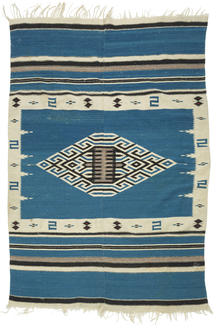 This serape was handwoven in central Mexico during the middle of the 20th century.

A luminous Texcoco serape distinguished by its saturated indigo field and a well articulated central diamond medallion. The stepped, labyrinthine geometry—woven in crisp natural white and deep black creates a powerful visual anchor, while the serrated motifs and butterfly-like accents along the border add nice rhythmic movement.