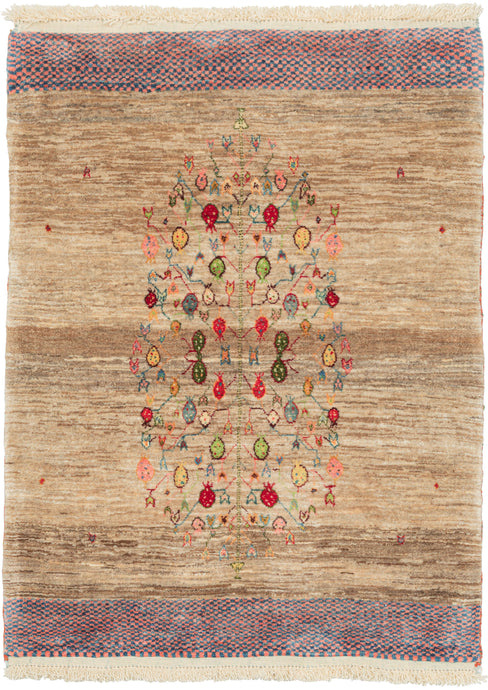 It features a a central tree of life motif with branches full of pomegranates in vibrant reds, blues, yellow and green. The tree sits atop a variegated field made up of various shades of undyed wool with a few small moments of color that add depth. Framed on top and bottom with a blue and pink checkered band. The pile is densely woven, making this a plush rug.