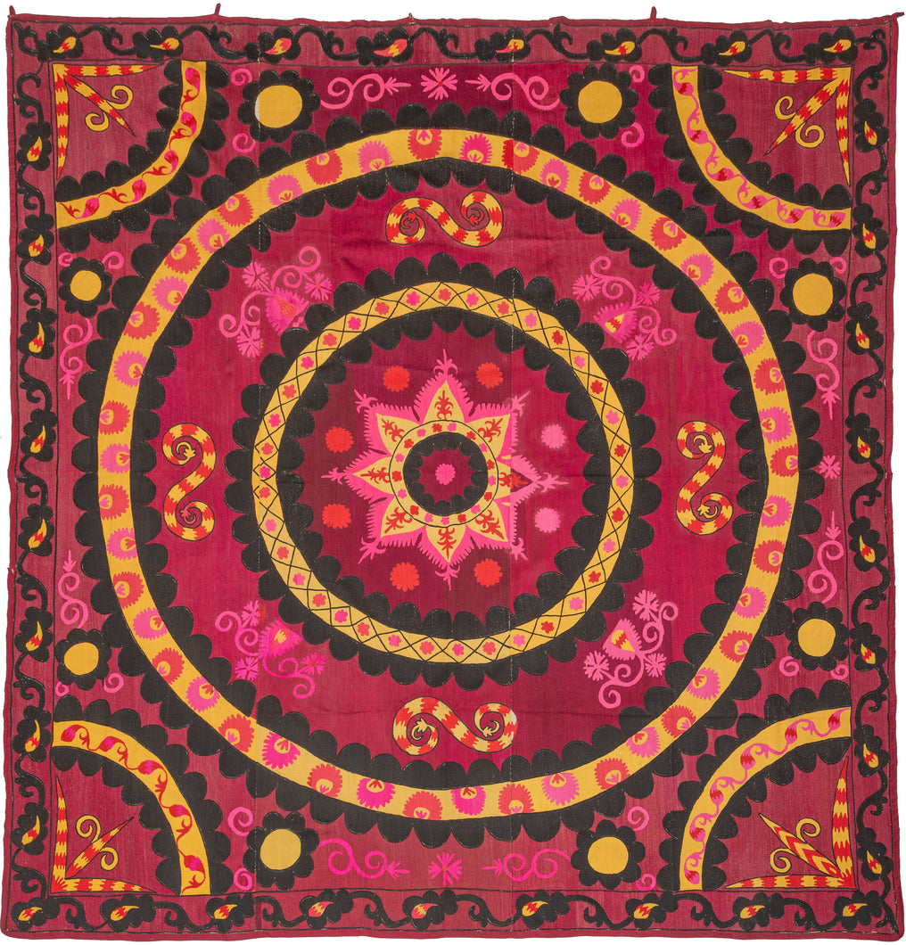 A bold and vibrant suzani centered around a large circular medallion radiating outward in layered rings of embroidery. The composition is energized by a rich crimson ground with striking accents of black, golden yellow, and hot pink, creating a dramatic and highly graphic presence.