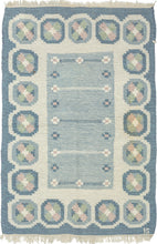 A refined mid-century Swedish flat weave by celebrated designer Ingegerd Silow, signed with her "IS" monogram in the lower right corner. Woven in soft glacier blues, cream, rose, and sage, this piece embodies the quiet elegance and warmth that define Scandinavian Modern textile design.

Silow’s signature geometric floral medallions create a rhythmic border around a subtly striated blue field, giving the rug both structure and softness. Handwoven in wool on a linen foundation, the texture is supple yet sturd