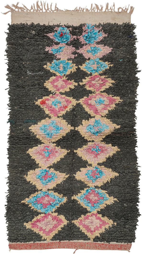 Boucherouite rugs, created by Amazigh (Berber) women in rural Morocco, transform recycled textiles into exuberant works of folk art. This example features a striking vertical column of lozenge motifs rendered in pink, sky blue, and pale yellow against a deep charcoal ground. The palette and high, shaggy pile give it a wonderfully tactile and expressive presence. A spontaneous composition with rhythmic repetition and playful color balance.
