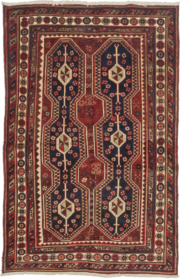 A Richly colored tribal rug with a striking double-column format of two tall elongated medallions&nbsp;on a deep indigo ground, each structured around a vertical spine of diamond forms quartered in olive and cream. The m&nbsp;red field between and around them reads almost as a third element, giving the composition an unusual density. Colors throughout are well-preserved with navy, red, ivory, and a muted green that surfaces in the medallion interiors and guard borders.