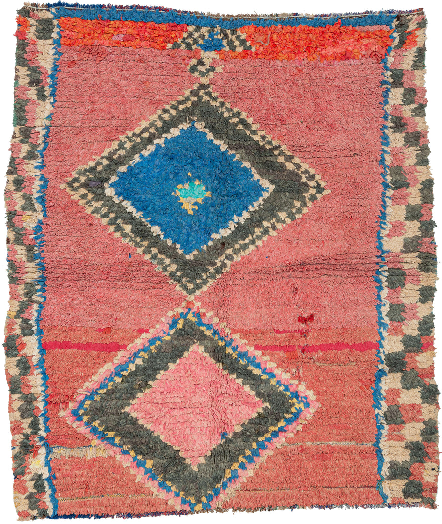 Vibrant and expressive, this Boucherouite rug embodies the joyful spontaneity of Moroccan village weaving. Crafted from recycled textiles, it features two large diamond medallions floating on a coral-pink ground, their layered outlines in blue, charcoal, ivory, and soft yellow creating a lively sense of rhythm and depth. A checkerboard border and flashes of crimson and turquoise add further energy to the composition.