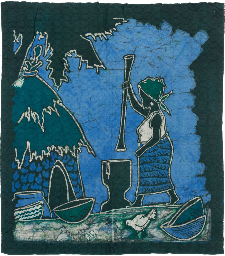 A striking West African batik textile worked in deep indigo, teal, and blue tones. The composition shows a standing figure using a tall pestle with a mortar, set within a stylized village landscape with a thatched structure, bowls, palms, and birds.